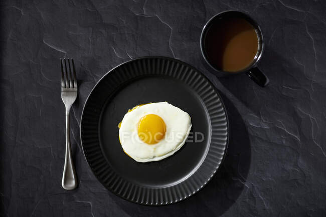 Sunnyside egg with fork and coffee on black surface with black plate and black coffee cup — Stock Photo