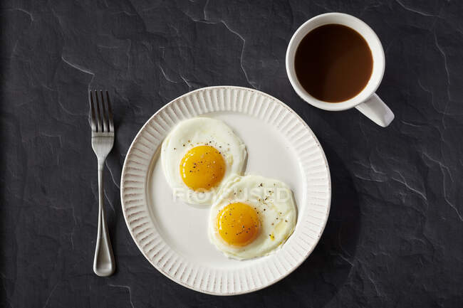 Two fried eggs with pepper in a white plate with coffee cup on black surface — Stock Photo