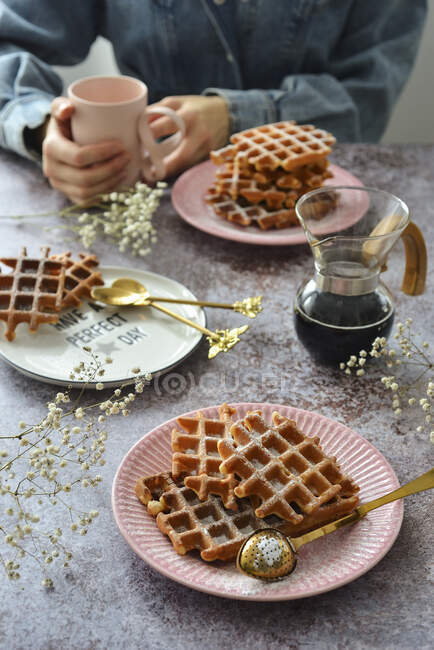 Waffles em um pires na mesa um jarro de café uma caneca. A mulher está segurando um copo em suas mãos — Fotografia de Stock