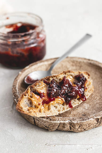 Bread slice with Peanut Butter and Jam on rustic clay plate with spoon and jar on background — Stock Photo