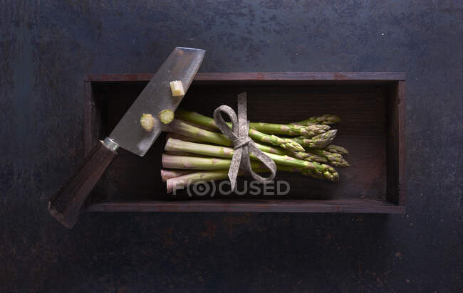 Un paquete de espárragos verdes frescos con un cuchillo en una caja de madera sobre una superficie oscura - foto de stock
