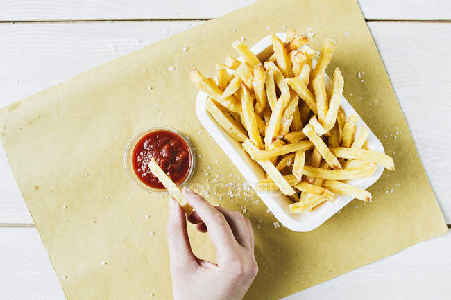 Una porción de papas fritas con sal marina, con una mano sumergiendo una en ketchup - foto de stock
