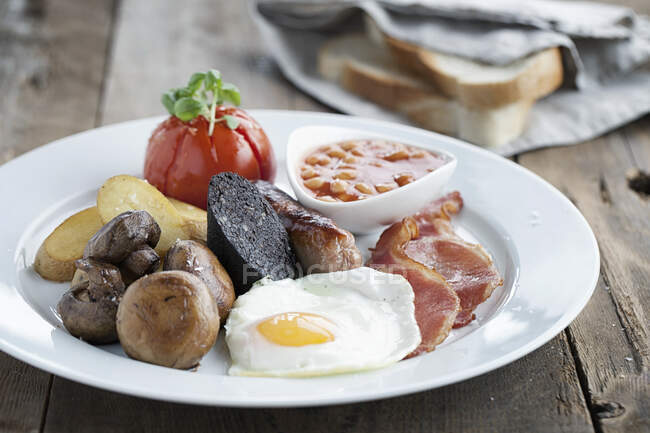 Английский завтрак подается на белой тарелке с белым нарезанным хлебом на заднем плане — стоковое фото