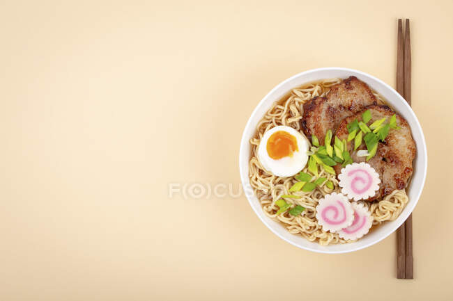 Верхній вигляд японського раму супу з локшини в білій мисці з локшиною, м'ясним бульйоном, нарізаною смаженою свининою, нарутомакі, яйце з жовткою на пастельному бежевому тлі. — стокове фото