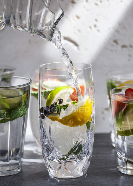 Vários copos de água cheios de frutas, hortelã e gengibre com água jorrando do jarro em um copo — Fotografia de Stock