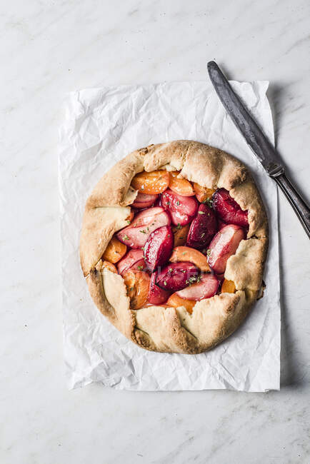 Galette pie filled with cut plums and knife on paper, top view — Stock Photo