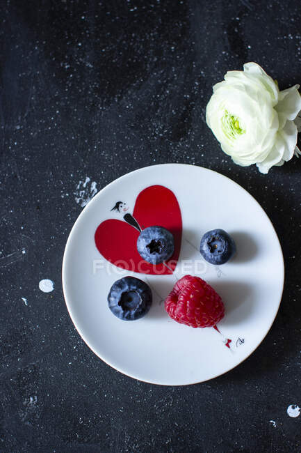 Framboesa fresca e mirtilos em pequeno prato com coração e flor na mesa — Fotografia de Stock