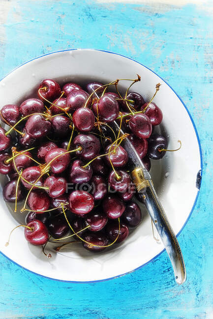 Fresh cherries in enamel bowl with knife on blue wooden surface — Stock Photo
