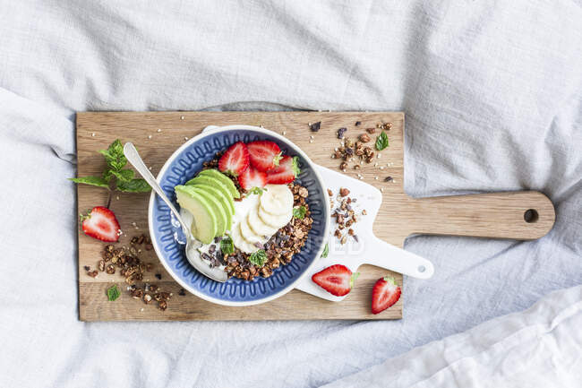 Almond and sesame seed granola with strawberries and avocado on wooden board on bed — Stock Photo