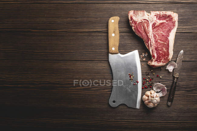 Un steak cru en T-bone avec des épices, un couteau et un fendoir sur une surface en bois — Photo de stock