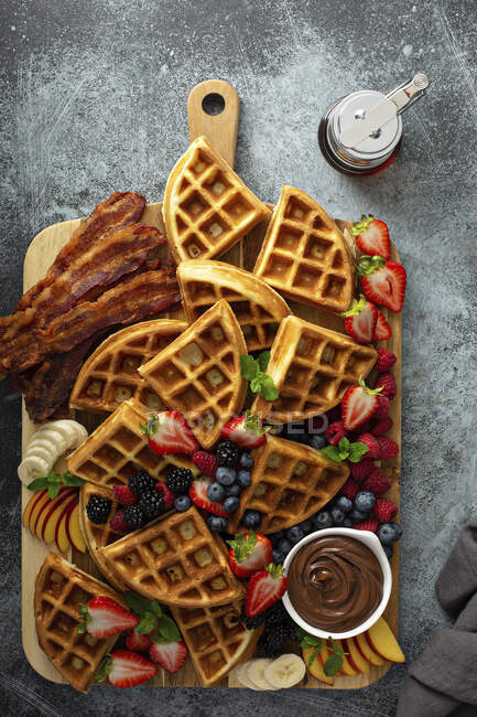 Большой вафельный бар на завтрак подается на деревянной доске с ягодами, беконом и шоколадным соусом — стоковое фото
