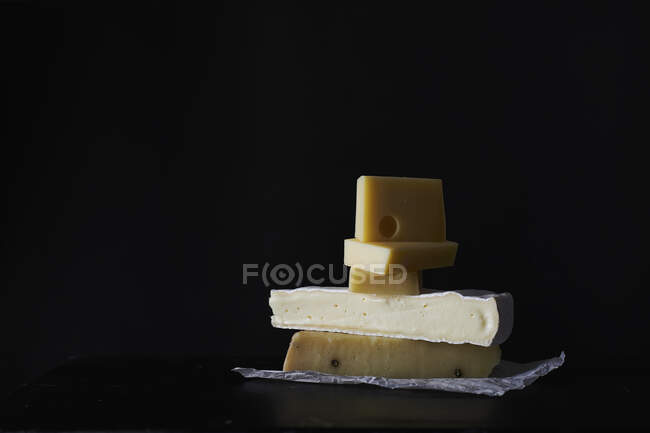 La pila de los pedazos distintos de queso sobre el papel al fondo oscuro - foto de stock