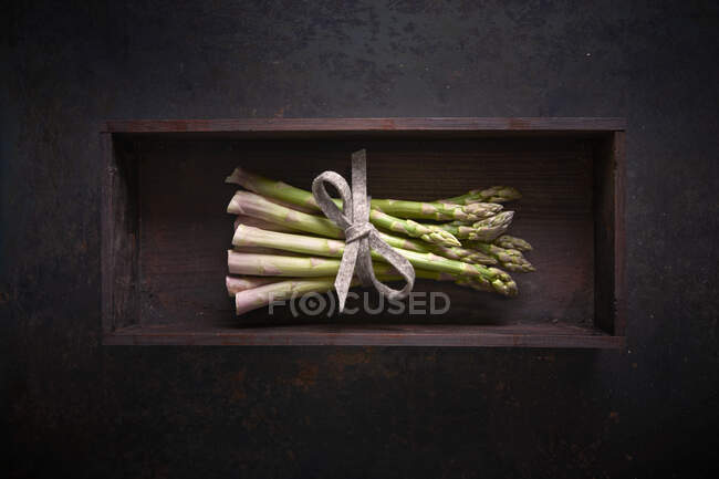 Un fascio di asparagi verdi freschi in una scatola di legno su una superficie scura — Foto stock