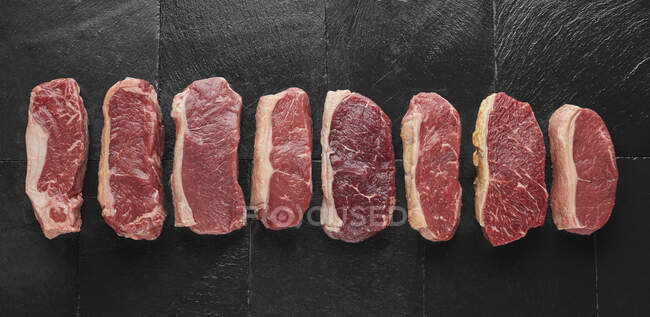 Mehrere rohe Steaks auf einem schwarzen Schieferteller — Stockfoto