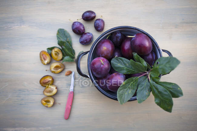Fresh plums in pot and halved with knife on wooden background — Stock Photo