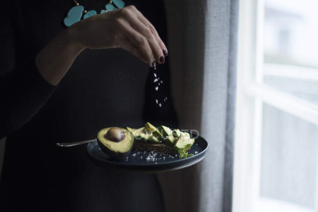 A woman standing at a window sprinkling fleur de sel onto avocado bread — Stock Photo