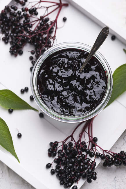 Black elderberries jam in glass jar with spoon surrounded with fresh berries and leaves — Stock Photo
