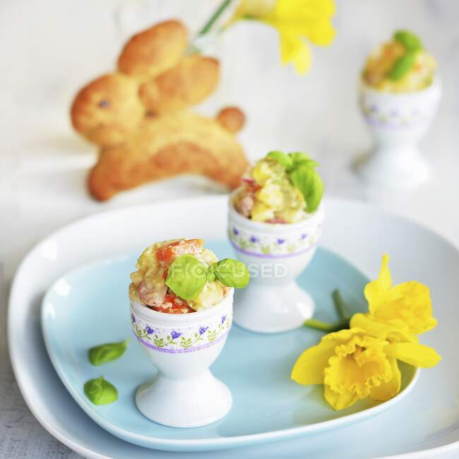 Potato salad with carrot, apple, peas and mayonnaise served in egg cups, with a yeast bread Easter bunny and daffodils in the background — Stock Photo