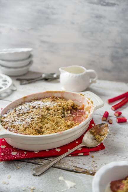 Rhubarb crumble in a baking dish with a jug of cream behind and fresh rhubarb — Stock Photo