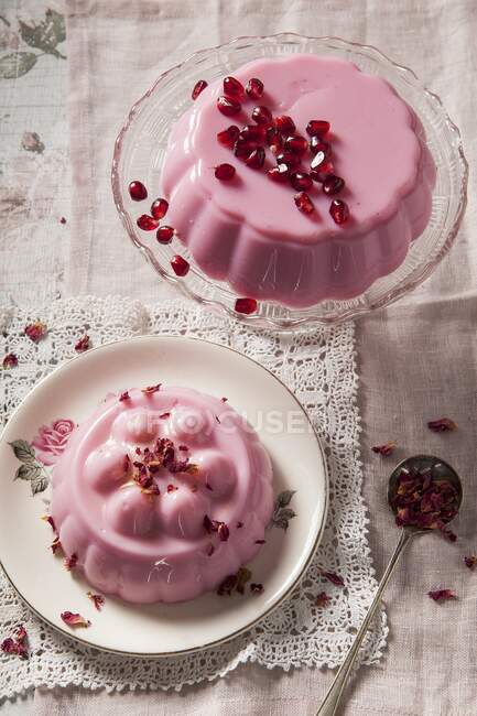 Два пастельних желе рожевого молока на старовинній скляній підставці і тарілці одна вкрита насінням грагранта, інша з сушеними пелюстками троянд, вистріленими над старовинною мереживною серветкою — стокове фото