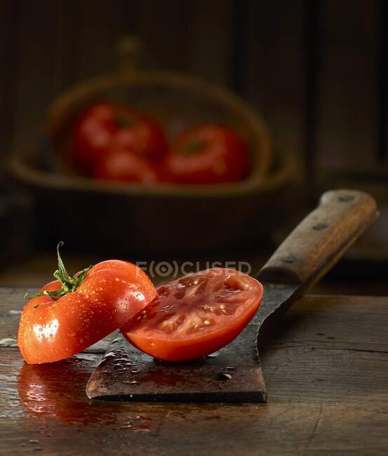 Un tomate a la mitad con gotitas de agua en un cuchillo de carnicero viejo - foto de stock