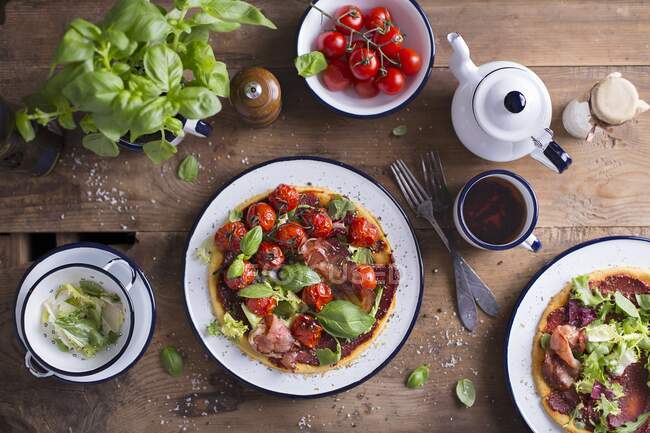 Pizza sin gluten con jamón de parma, hojas de albahaca fresca y tomates cherry servidos en un plato de esmalte, en una mesa de madera - foto de stock