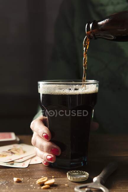 Botella de Guinness se vierte en un vaso grande que se sostiene por una mano con esmalte de uñas rojo en una mesa de madera rodeada por la tapa de la botella, abridor de botellas, nueces y cartas de juego - foto de stock