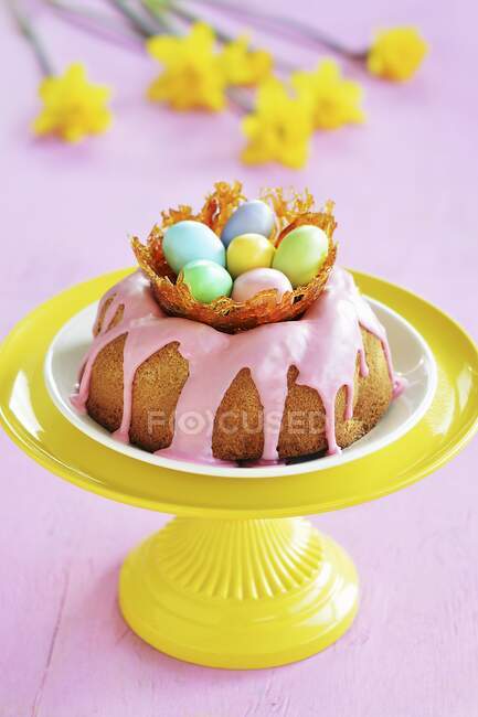 Ein Gugelhupf mit rosa Zuckerglasur und ein Karamellnest mit bunten Zuckereiern auf einem Ständer, im Hintergrund Narzissen — Stockfoto