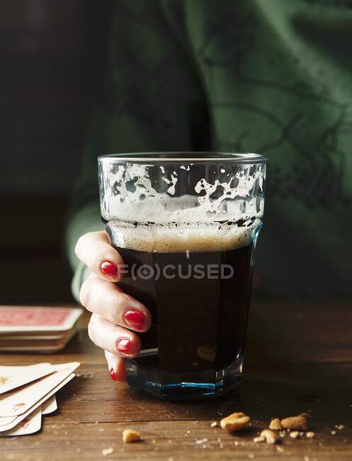 Copa de Guinness borracho con una mano de damas con clavos rojos y un suéter verde sosteniendo vidrio en una mesa de madera rodeada de nueces y jugando a las cartas - foto de stock