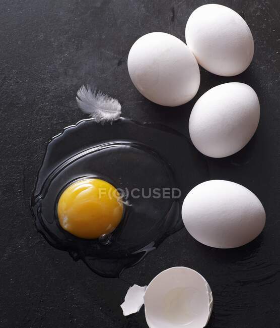 Ovos inteiros e ovos líquidos crus com casca quebrada e penas na superfície preta — Fotografia de Stock