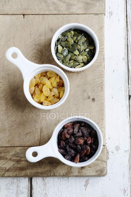 Raisins, golden raisins and pumpkin seeds in white bowls, seen from overhead — Stock Photo