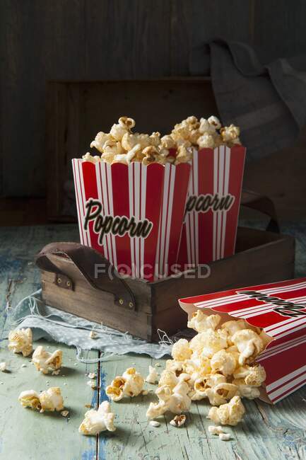 Two red and white striped boxes of popcorn on a wooden tray one box tipped over with popcorn spilling out onto an aqua green blue wooden surface - foto de stock