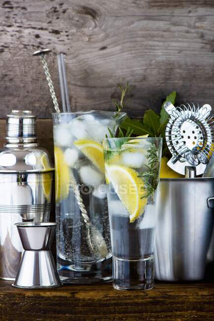 Gin and tonic with lemon, ice cubes and rosemary between various bar utensils — Stock Photo