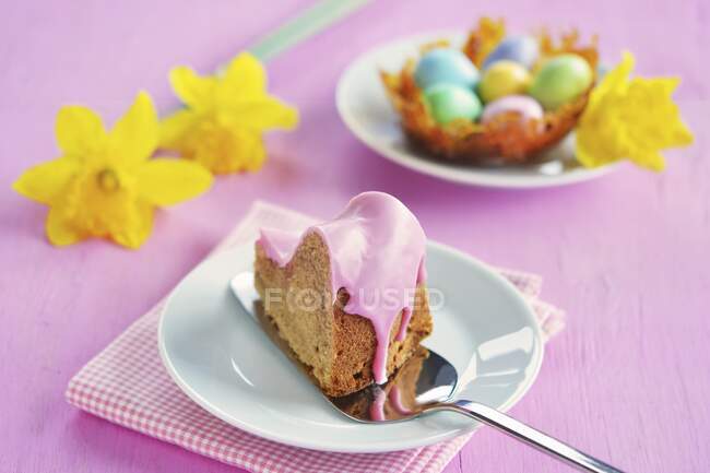 Кусок гугельхупфа с розовой сахарной глазурью, карамельное гнездо с красочными сахарными яйцами и нарциссами на заднем плане — стоковое фото