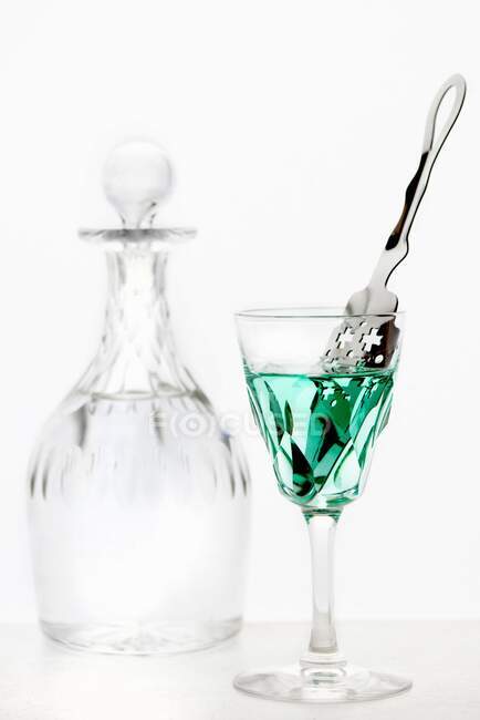 Absenta con una cuchara de absenta en un vaso de cristal frente a una jarra de cristal de agua - foto de stock