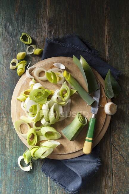 Foto aérea de puerros picados y preparados en una tabla redonda de cortar de madera por un cuchillo de mango verde y amarillo sobre un paño marino y una superficie de madera verde - foto de stock