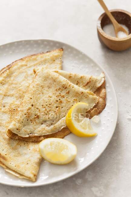 Two folded pancakes on an off white texture plate with lemon wedges on a stone surface — Stock Photo