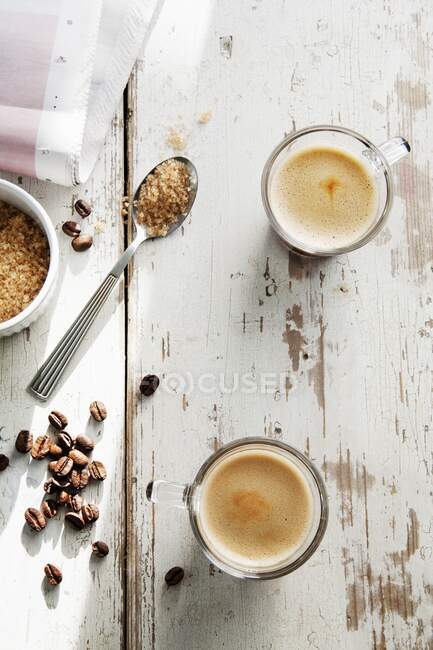 Granos de café, una cucharadita de azúcar morena y dos tazas de espresso en una mesa blanqueada - foto de stock