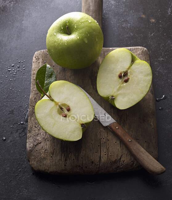 Manzanas Granny Smith, enteras y cortadas a la mitad, sobre una vieja tabla de madera con un cuchillo - foto de stock