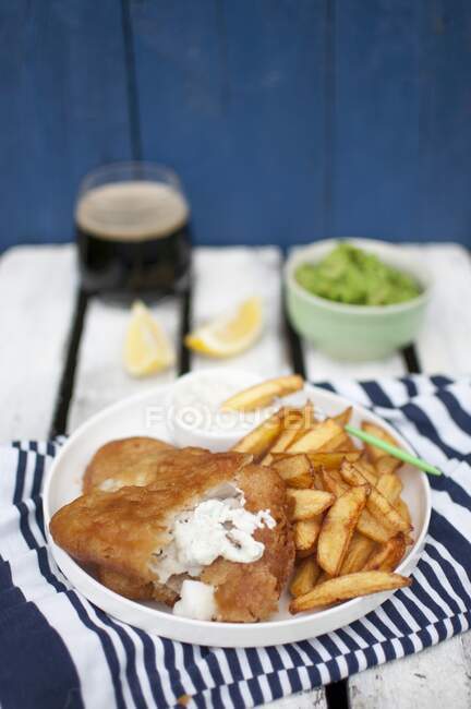 Peixe e batatas fritas, servidos com purê de ervilhas verdes, pedaços de molho de limão tártaro e cerveja escura — Fotografia de Stock