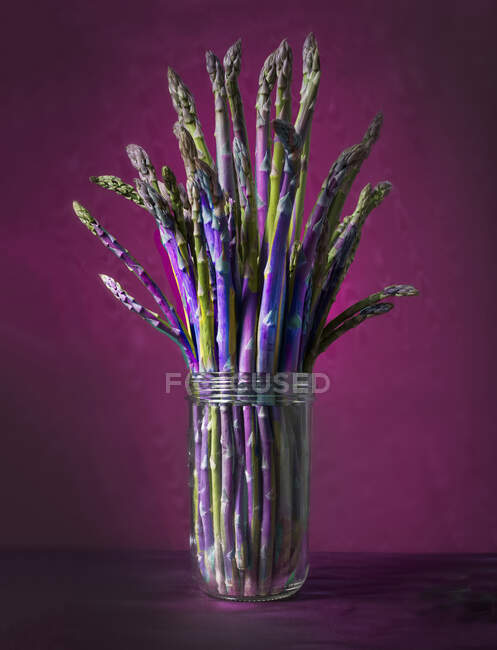 Lances d'asperges fraîches dans un verre sur fond rouge-violet — Photo de stock