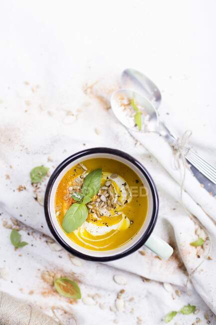 Гарбузовий суп в емальованому кухоль; прикрашений свіжим базиліком і насінням соняшнику — стокове фото