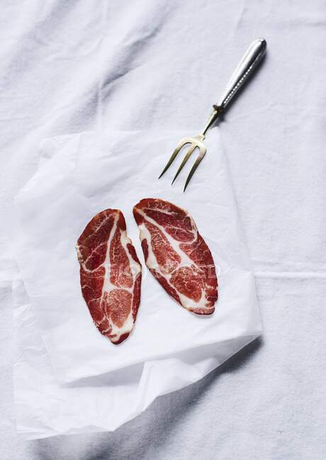 Dos rebanadas de carne curada con un tenedor de carne sobre una hoja de papel - foto de stock