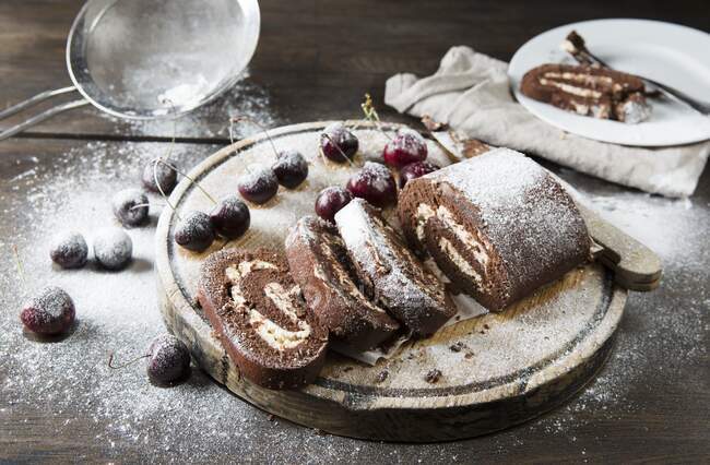 Chocolate swiss roll whole and chopped into slices with cherries and icing sugar — Stock Photo