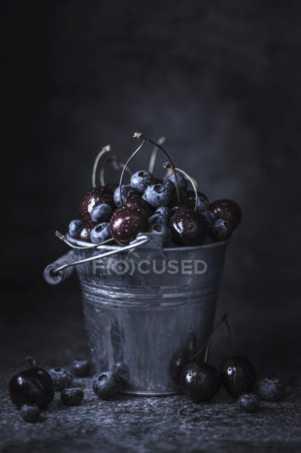 Mirtilos e cerejas em mini balde de metal e na superfície da mesa — Fotografia de Stock