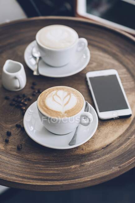 Dos tazas de capuchino junto a un smartphone sobre una mesa en un café - foto de stock