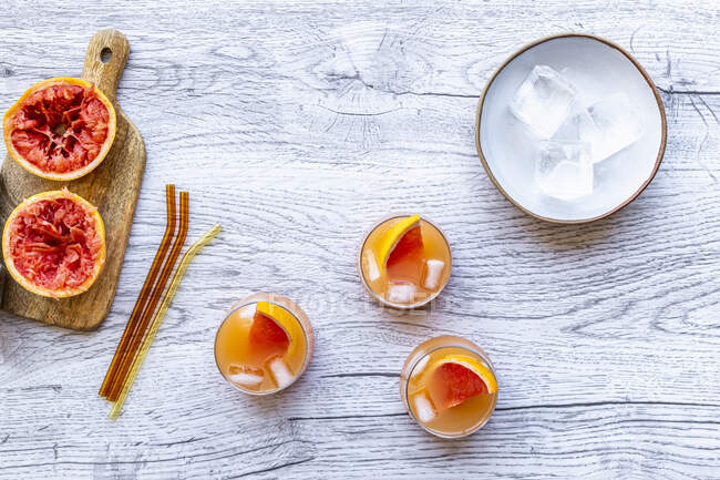 Cocktail di pompelmo e vodka in bicchieri su superficie di legno con cannucce, ghiaccio e bucce di pompelmo — Foto stock