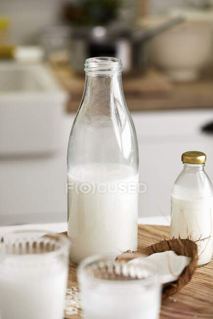 Молоко, бутылки и овсяные зёрна в стеклянных банках и кувшин на деревянном столе — стоковое фото