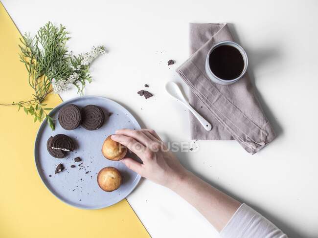 Uma caneca de café ao lado de uma mão tirando um bolo de um prato — Fotografia de Stock