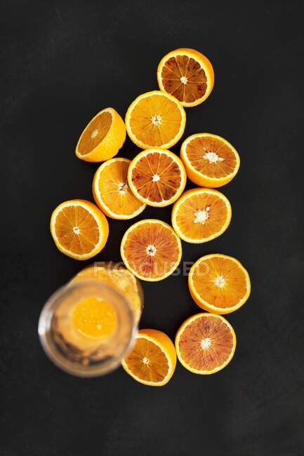 A glass jug of orange juice and halved Moro oranges against a black background — стокове фото